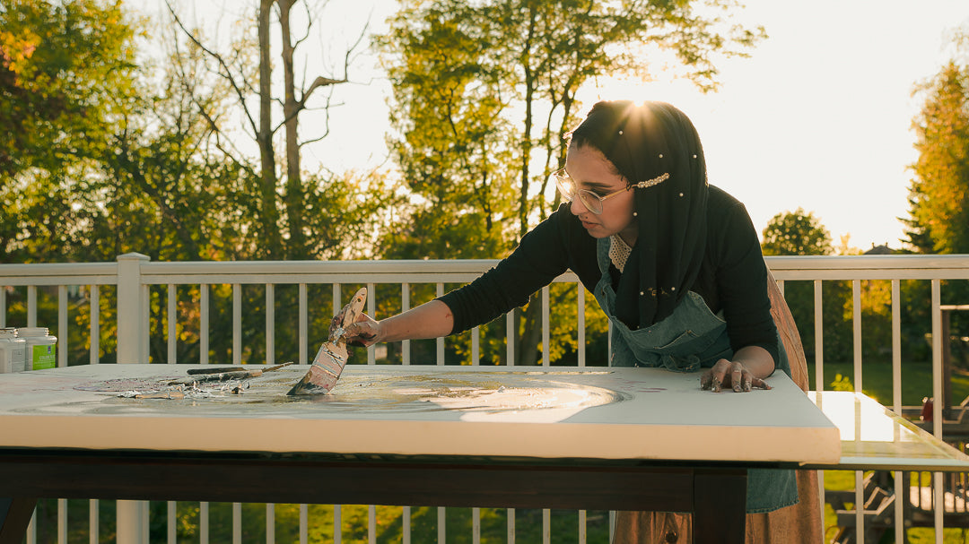 A woman wearing a black hijab and denim apron paints on a large white canvas laid out on a table on a sunny outdoor deck with trees in the background.