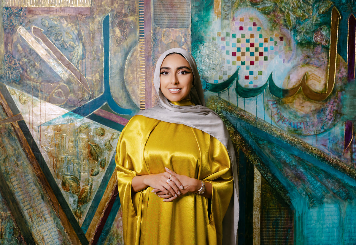 A woman wearing a shiny mustard yellow abaya and a light gray hijab stands in front of a large, colorful Islamic calligraphy painting.